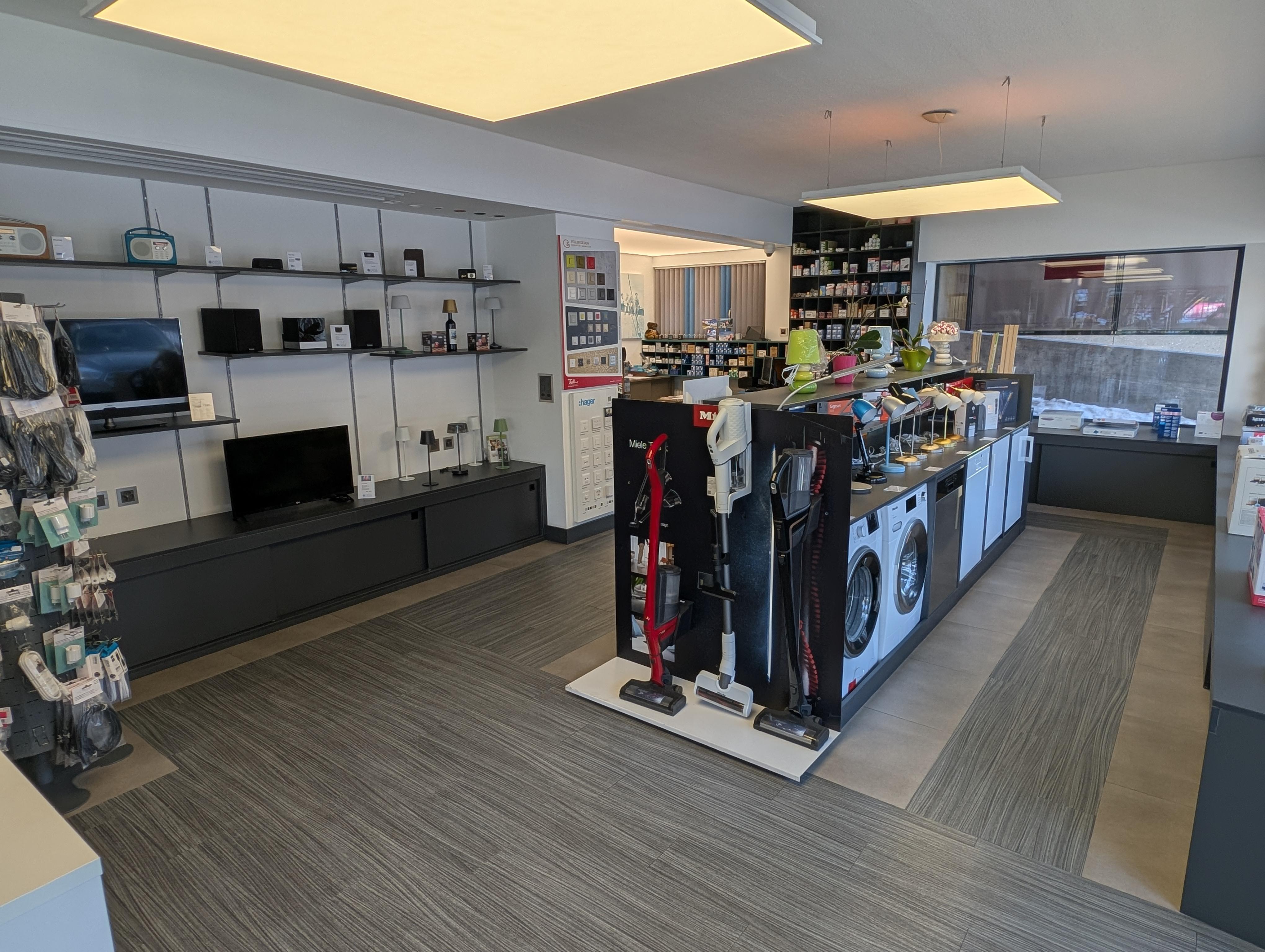 Présentoir dans un showroom d'électroménager avec machines à café Nespresso et appareils Miele, éclairage chaleureux et cadre fin rappelant l'identité artisanale valaisanne